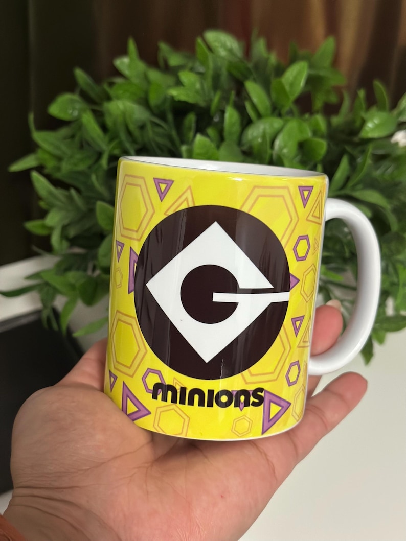 May include: A yellow ceramic mug featuring the Minions logo and the word "minions" in black. The mug has a white handle and is decorated with purple triangles and yellow hexagons. The mug is held in a hand.