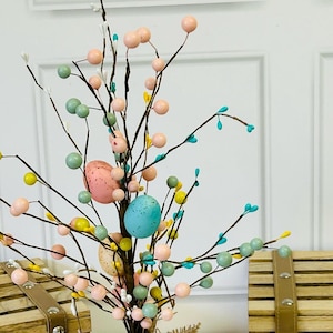 May include: A small Easter tree with brown branches decorated with pastel pink, blue, and yellow eggs and white, pink, and green berries. The tree is in a burlap bag.