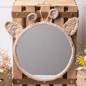 Puede incluir: Un espejo redondo con un marco de crochet beige con forma de cara de animal con dos orejas. El espejo está colgado en una caja de madera con una correa de cuero marrón.