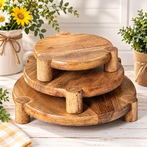 May include: A set of three round, light brown wooden risers stacked on top of each other. Each circular riser features a smooth, natural wood grain finish and is supported by three short, cylindrical legs, creating a tiered display structure.