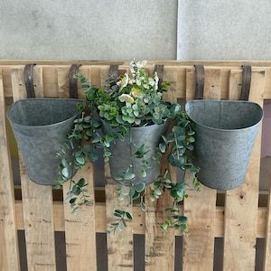 May include: Three galvanised metal hanging planters with green plants and white flowers, hanging on a wooden pallet wall.