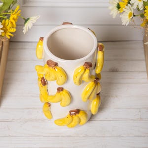 Peut inclure: Un vase en c&eacute;ramique blanc, de forme cylindrique, orn&eacute; de motifs en forme de bananes jaunes. Les bananes ont des tiges marron et sont dispos&eacute;es autour du vase. Le vase est vide et pos&eacute; sur une surface en bois blanc.