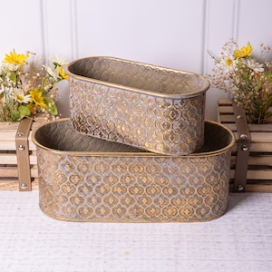 Può includere: Due vasi ovali in metallo con finitura oro e bianco invecchiato. I vasi hanno un motivo decorativo e sono impilati uno sopra l'altro.