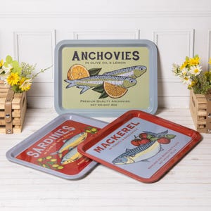 May include: Three rectangular metal trays featuring vintage-style illustrations of fish. One tray is light blue with anchovies and lemon, one is grey with sardines and chilli, and one is red with mackerel and tomatoes. Each tray lists net weight 4oz.