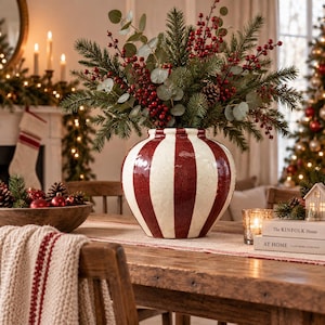 May include: A round ceramic vase featuring vertical stripes in deep red and off-white. The vase is filled with a festive arrangement of green pine branches, eucalyptus leaves, and clusters of small red berries, creating a holiday-themed centrepiece.