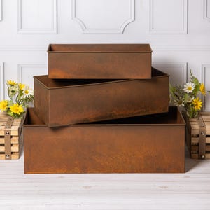 May include: A set of three rectangular metal planters with a weathered, rusted brown finish. The containers are stacked in a tiered arrangement, featuring clean, straight edges and an open top design, showcasing a consistent oxidised metal texture.