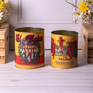 Może przedstawiać: Dwa cylindryczne metalowe pojemniki z żółtymi i czerwonymi etykietami. Jeden z napisem "El Toro Smoked Paprika Caliente", drugi "Las Palmas Spanish Red Chili Sauce". Oba mają wygląd vintage, prawdopodobnie do kuchni.