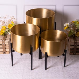 Op de afbeelding: Drie goudkleurige metalen plantenbakken van verschillende formaten. Elke plantenbak heeft een ronde komvorm en staat op drie zwarte metalen poten. Geschikt voor binnen- of buitendecoratie.