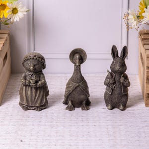 May include: Three detailed figurines: a hedgehog holding a teacup, a goose wearing a bonnet, and a rabbit holding a carrot. The figurines are bronze-colored and stand on a light-colored surface. The theme is reminiscent of a children's story.