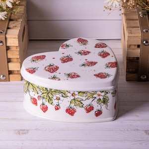 Peut inclure: Une boîte à bijoux en céramique en forme de cœur, blanche avec un couvercle orné de fraises rouges. Les côtés présentent un motif de vigne de fraises avec des feuilles vertes, des fleurs blanches et des fraises rouges.