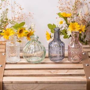 May include: Four decorative glass bud vases in clear, green, gray, and pink. The vases have various textures and shapes, including ribbed and patterned designs. They are arranged on a wooden crate with floral accents in the background.