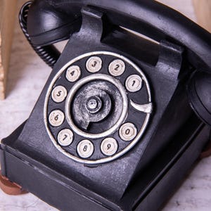 May include: A black rotary phone with a silver dial and white numbers. The phone has a vintage design and is made of plastic.