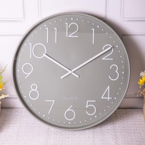 May include: A large, round, grey wall clock with white numbers and hands. The clock face has a subtle texture and the words "QUARTZ" are printed in white at the bottom.