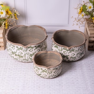 May include: A trio of ceramic planters with a heart-shaped rim. The planters have a white base with a green, leafy scroll design. The rims are light brown, and the interiors are a muted grey colour.