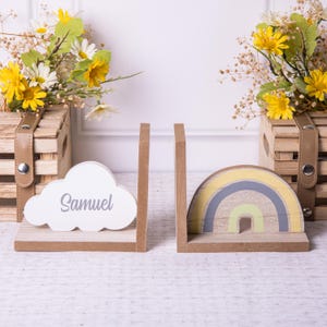 May include: A set of wooden bookends featuring a rainbow and a cloud with the name Samuel. The bookends are painted in yellow, gray, green, and white.