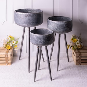 May include: Three galvanised metal planters with a distressed finish. Each planter has a rounded bowl shape and sits on three slender, pointed legs. The planters are different sizes, creating a tiered display. The background includes flowers and wooden crates.