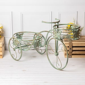 May include: A decorative, antique-style metal tricycle planter in a distressed, light green finish. The tricycle features two round baskets and butterfly accents. The wheels and frame have a scrolled design. The planter is designed to hold flowers or plants.