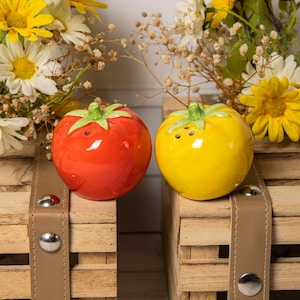 Peut inclure: Une paire de salières et poivrières en céramique en forme de tomates. L'une est rouge avec une tige verte, l'autre est jaune avec une tige verte. Elles sont posées sur une caisse en bois avec une sangle en cuir.