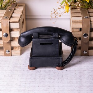May include: A black rotary phone shaped piggy bank with a coiled cord. The phone has a slot for coins on the side. The piggy bank is sitting on a wooden surface.
