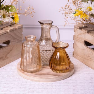 Op de afbeelding: Drie glazen vazen in verschillende vormen en maten op een ronde houten basis. De vazen zijn helder, amberkleurig en lichtbruin. De vazen zijn leeg.