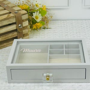 May include: White jewellery box with a clear lid and a drawer. The lid has a compartment for rings and other jewellery. The drawer has a clear acrylic knob. The box is personalised with the name "Maura".