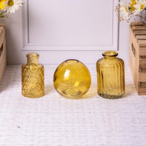 Op de afbeelding: Drie amberkleurige glazen vazen in verschillende vormen. De vaas links is rechthoekig met een ruitpatroon. De middelste vaas is bolvormig en de rechter vaas is cilindrisch met verticale ribbels. Decoratieve woondecoratie.