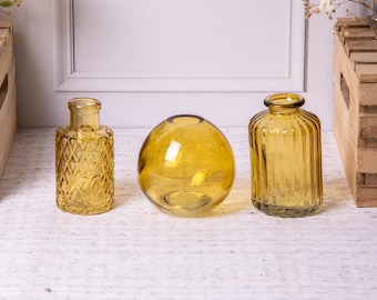 Könnte beinhalten: Drei bernsteinfarbene Glasvasen in verschiedenen Formen. Die linke Vase ist rechteckig mit einem Rautenmuster. Die mittlere Vase ist kugelförmig und die rechte Vase zylindrisch mit vertikalen Rillen. Dekorative Wohnkultur.
