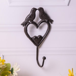 May include: A dark brown metal wall hook featuring two birds kissing above a heart-shaped design. The hook has a small heart at the bottom of the larger heart and a curved hook for hanging items. The wall hook is mounted on a white wall.