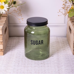 May include: A cylindrical olive green glass storage jar with a black metal lid. The word SUGAR is printed in black sans-serif capital letters on the front of the jar. The glass is translucent, allowing for a view of the interior space.