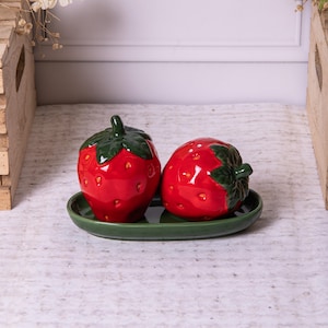 May include: A ceramic salt and pepper shaker set shaped like strawberries. The red shakers have green leafy tops and are on a green oval tray. The set is designed to resemble the fruit.