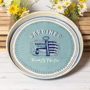 Könnte beinhalten: Ein rundes Metalltablett mit abgenutztem hellblauem Finish, das in der Mitte eine Sardinendose mit dem Text SARDINES, JUST TAKE A BITE, SARDINES CLUB und Fruit Of The Sea zeigt. Das Tablett hat dunkelblaue kreisförmige Randlinien.
