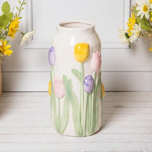 Może przedstawiać: Kremowy ceramiczny wazon z wytłoczonymi tulipanami w odcieniach żółci, różu i fioletu. Wazon ma cylindryczny kształt i zielone liście. Dekoracyjny przedmiot do wystroju domu.