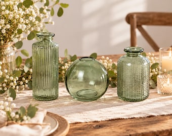 May include: Three sage green glass vases of varying shapes. The left vase is tall and cylindrical with vertical ribbing. The center vase is spherical and smooth. The right vase is shorter and wider with a textured grid pattern on the surface.