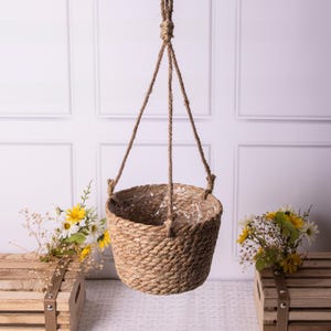 May include: A hanging woven basket made from natural fibres, suspended by a thick rope. The basket has a round shape and is designed for plants. The background is a white wall with decorative panelling. Flowers are placed on wooden crates.