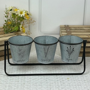 May include: Three galvanised metal planters with a black metal stand. The planters are labelled "parsley", "rosemary", and "thyme".