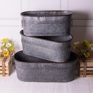 May include: Three grey metal oval planters with brown rims and handles. The planters are stacked on top of each other.