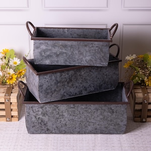 May include: Three rectangular galvanized metal planters with brown trim and handles are stacked. The planters have a rustic, textured appearance. Yellow and white flowers in wooden crates are on either side.