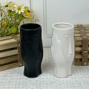 May include: Two ceramic vases with a face and hands design. One vase is black and the other is white.