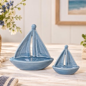May include: Two glossy, light blue ceramic sailboat figurines of different sizes. Each boat features a smooth, curved hull and a single mast with two triangular sails. The ceramic surface has a reflective finish, highlighting the blue color.