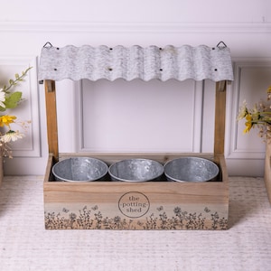 Könnte beinhalten: Ein Pflanzkasten aus Holz mit drei Metalleimern. Der Pflanzkasten hat ein Dach aus gewelltem Metall und ist mit einem Blumen- und Schmetterlingsdesign verziert. Der Text "the potting shed" ist auf der Vorderseite zu sehen.