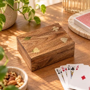 May include: A rectangular wooden box with a natural grain finish, featuring four inlaid brass suit symbols on the lid representing hearts, diamonds, clubs, and spades. The box has a hinged lid and a smooth, polished surface with visible wood texture.