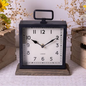May include: Black square clock with a white face and black hands. The clock has a metal frame and a wooden base. The clock face has black Roman numerals and the word "LONDON" printed below the 12.