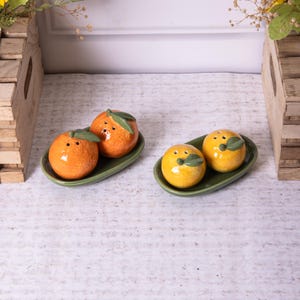May include: Two sets of ceramic salt and pepper shakers shaped like oranges. One set is orange and the other is yellow. The shakers are on green oval-shaped dishes.