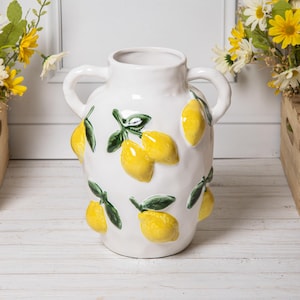 May include: White ceramic vase with two handles, adorned with raised yellow lemons and green leaves. The vase has a textured surface and a wide opening. A decorative piece for the home.