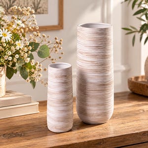 Può includere: Due vasi cilindrici con un motivo a venature del legno orizzontali in bianco e marrone chiaro. I vasi hanno una base liscia e arrotondata e una semplice apertura superiore. Il vaso più alto si trova a destra di quello più basso, entrambi coordinati.