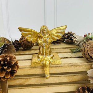 Puede incluir: Porta calcetines de ángel dorado con base y alas blancas. El ángel está arrodillado con las manos juntas en oración. El soporte está sobre una superficie de madera con piñas.