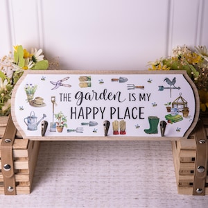 May include: A wooden sign with a white background and a green, brown, and black illustration of gardening tools and a garden scene. The sign reads "The garden is my happy place".