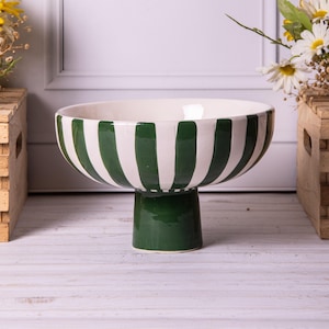Könnte beinhalten: Eine Keramik-Standfußschale mit weißem Innenraum und grün-weiß gestreiftem Äußeren. Die Schale steht auf einem einfarbigen grünen Sockel. Die Schale ist ein dekoratives Stück.