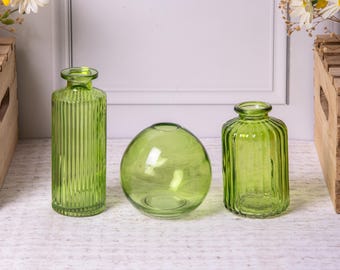 May include: Three green glass vases of varying shapes. The vase on the left is tall and cylindrical with vertical ridges. The center vase is spherical. The vase on the right is short and cylindrical with vertical ridges. The vases are on a white surface.
