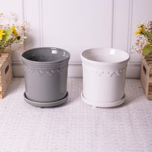 May include: Two ceramic plant pots with matching saucers. The pot on the left is grey and the pot on the right is white. Both pots have a scalloped design around the top.